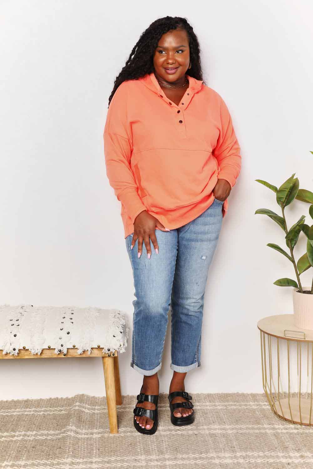 A woman with long hair wears a Double Take Quarter-Snap Dropped Shoulder Hoodie in solid orange. She is standing, with one hand touching her hair and the other resting in the pocket of her jeans, exuding a casual vibe.