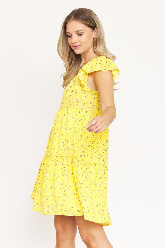 A woman wearing a Floral Flutter Sleeve Tunic with short ruffled sleeves stands against a plain background, smiling and holding a strand of her long, blonde hair.