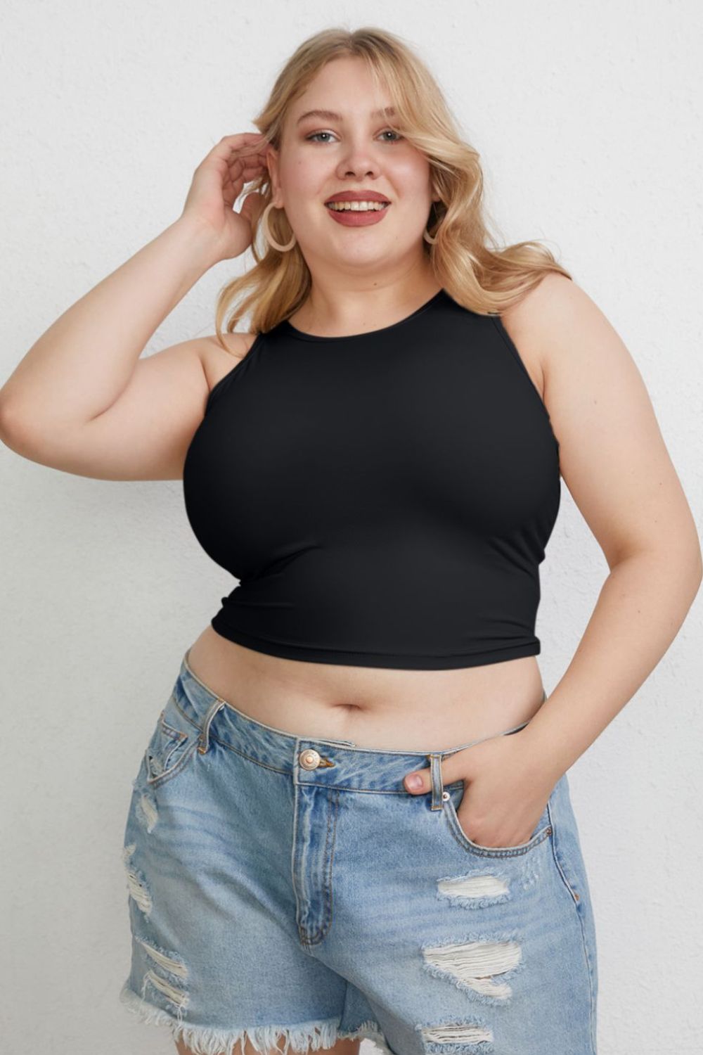 A woman with light hair wears the Basic Bae Full Size Round Neck Slim Tank in black and blue jeans, standing against a light-colored curtain.