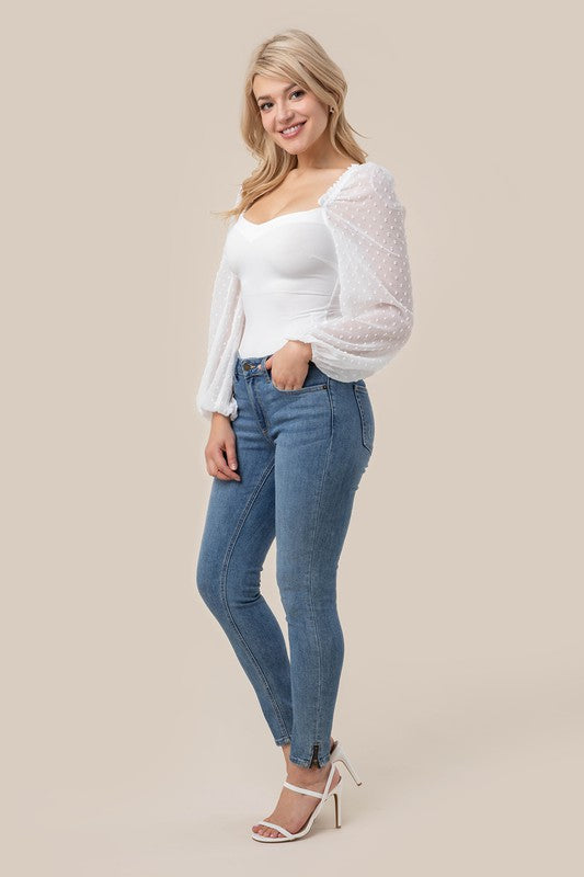 A person wearing the LS shirring sleeve bodysuit with Swiss dot mesh, featuring sheer, long puffed sleeves and a sweetheart neck, paired with light-colored jeans, stands against a plain background.