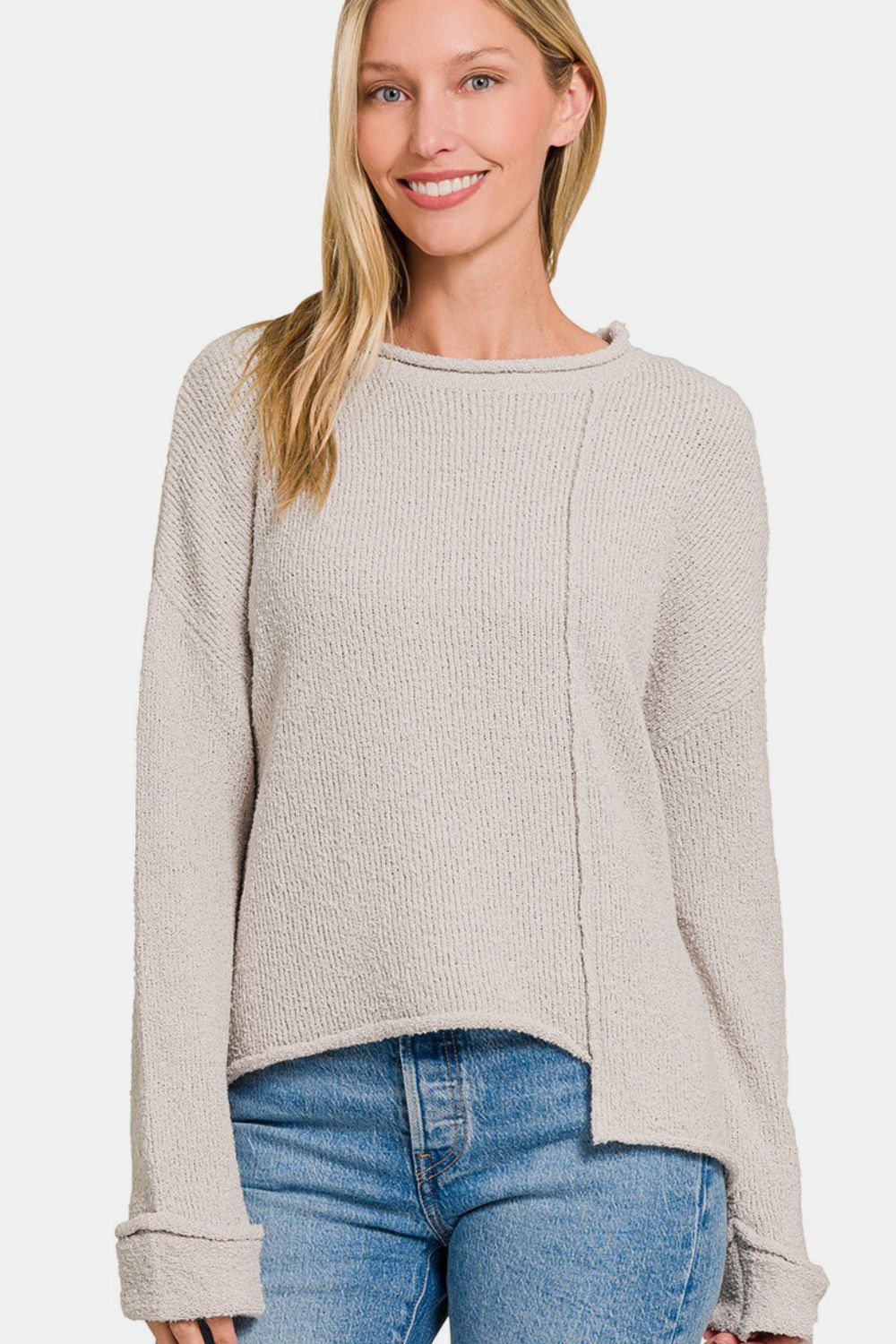 A woman in a light beige Zenana Asymmetric Hem Drop Shoulder Sweater poses with a hand near her head and another in her pocket, smiling slightly. She is wearing blue jeans against a plain white background.