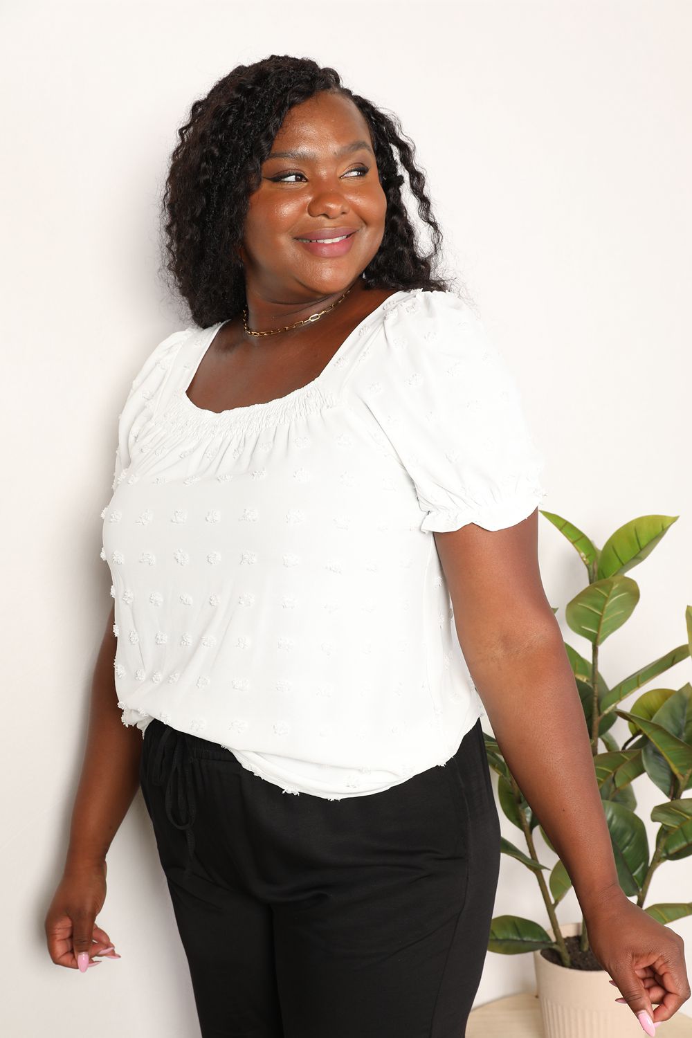 A person wearing the Mandy Swiss Dot Puff Sleeve Square Neck Blouse in white, paired with black pants, stands in front of a light background with green leaves visible on the right side.