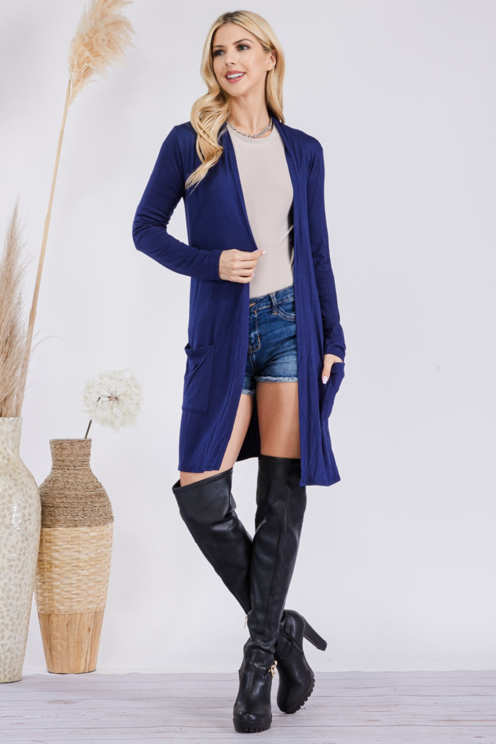 A woman stands indoors wearing the Celeste Full Size Open Front Cardigan with Pockets, a long blue USA-made cardigan. She pairs it with a beige top, denim shorts, and black thigh-high boots. She is smiling and looking to her left, with tall vases of dried plants beside her.