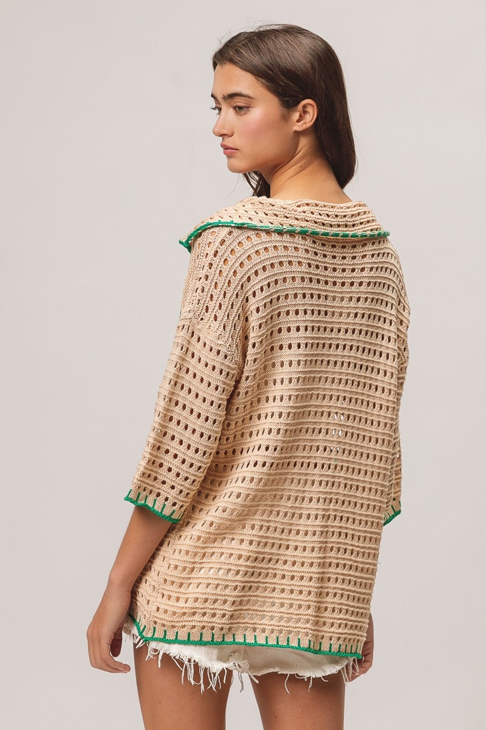 A woman wearing the BiBi Edge Stitched Cactus Patch Sweater Cardigan, featuring beige fabric with green cactus designs, layered over a white top and paired with white frayed shorts, stands against a plain background.