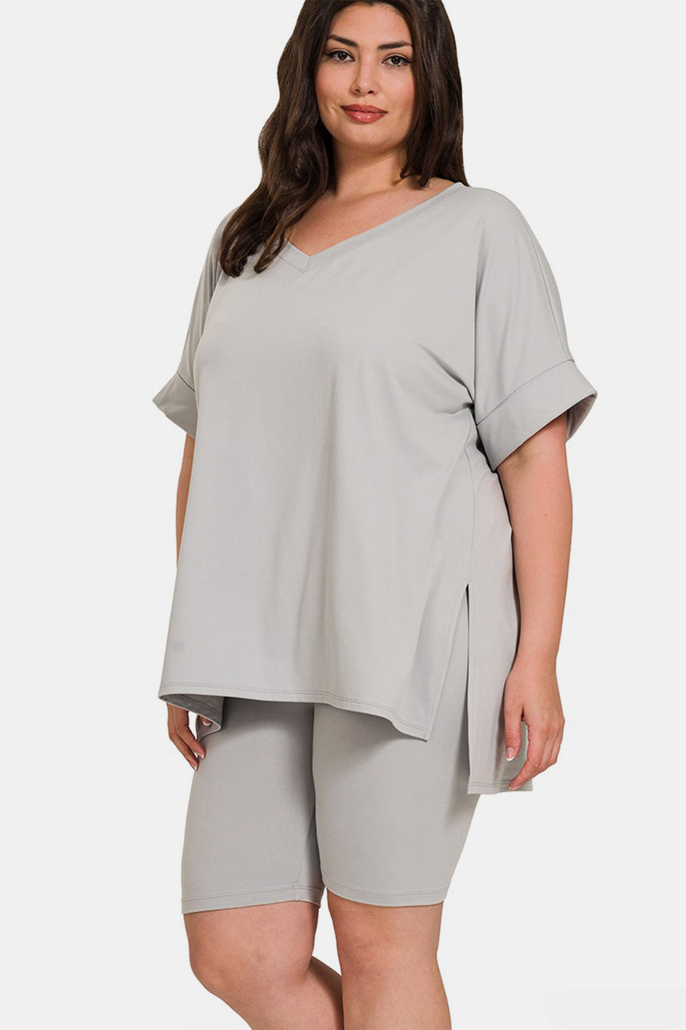 A woman is wearing the Zenana Full Size V-Neck Short Sleeve Slit T-Shirt and Shorts Set in a light gray color. The loose-fitting, highly stretchy outfit features a V-neck t-shirt with short sleeves and matching knee-length shorts. She is smiling with her hand raised near her head.