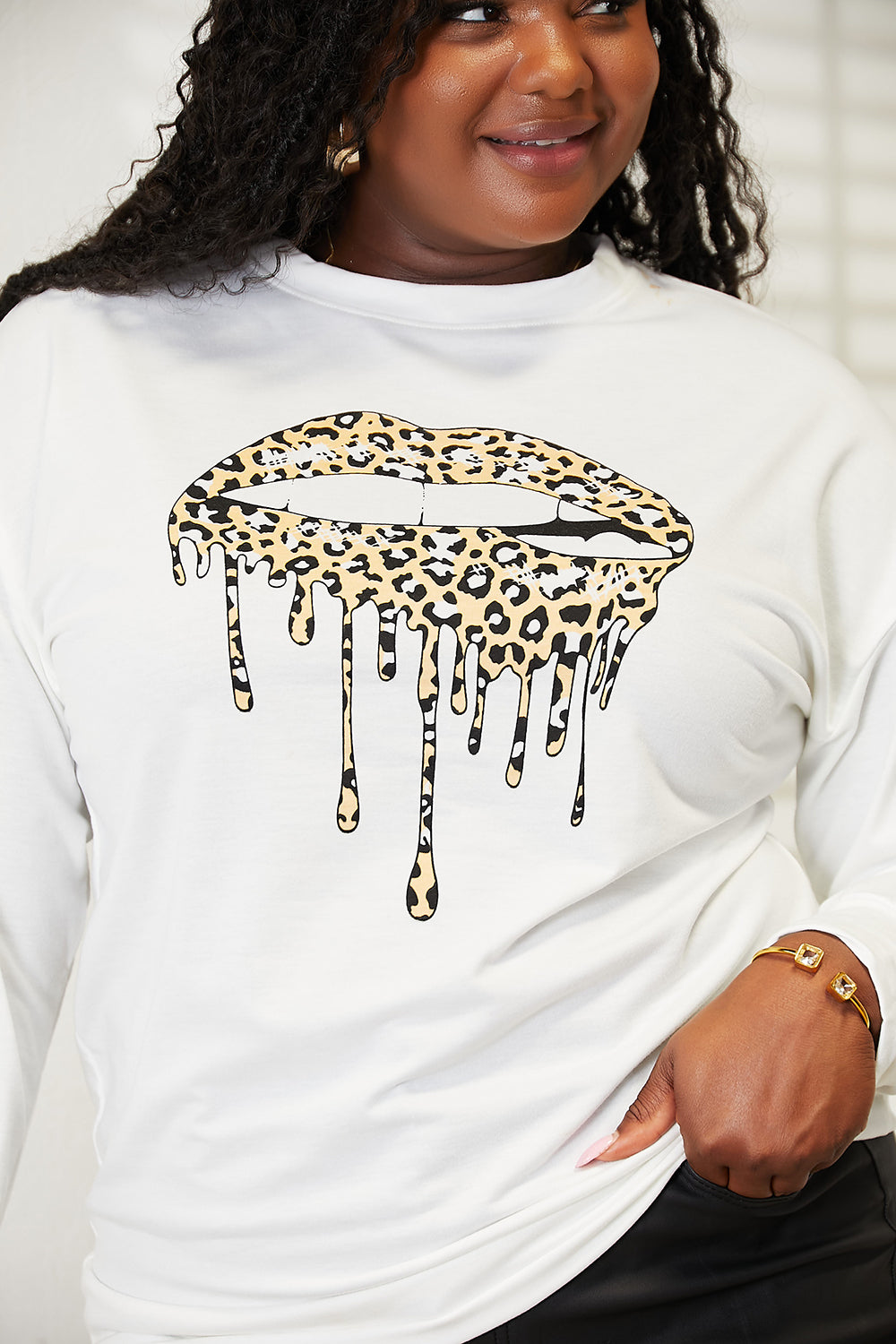 A woman stands indoors wearing a trendy and comfortable outfit—a white Simply Love Graphic Dropped Shoulder Round Neck Sweatshirt, featuring a dripping lip design, paired with black pants. One hand rests on a piece of furniture.