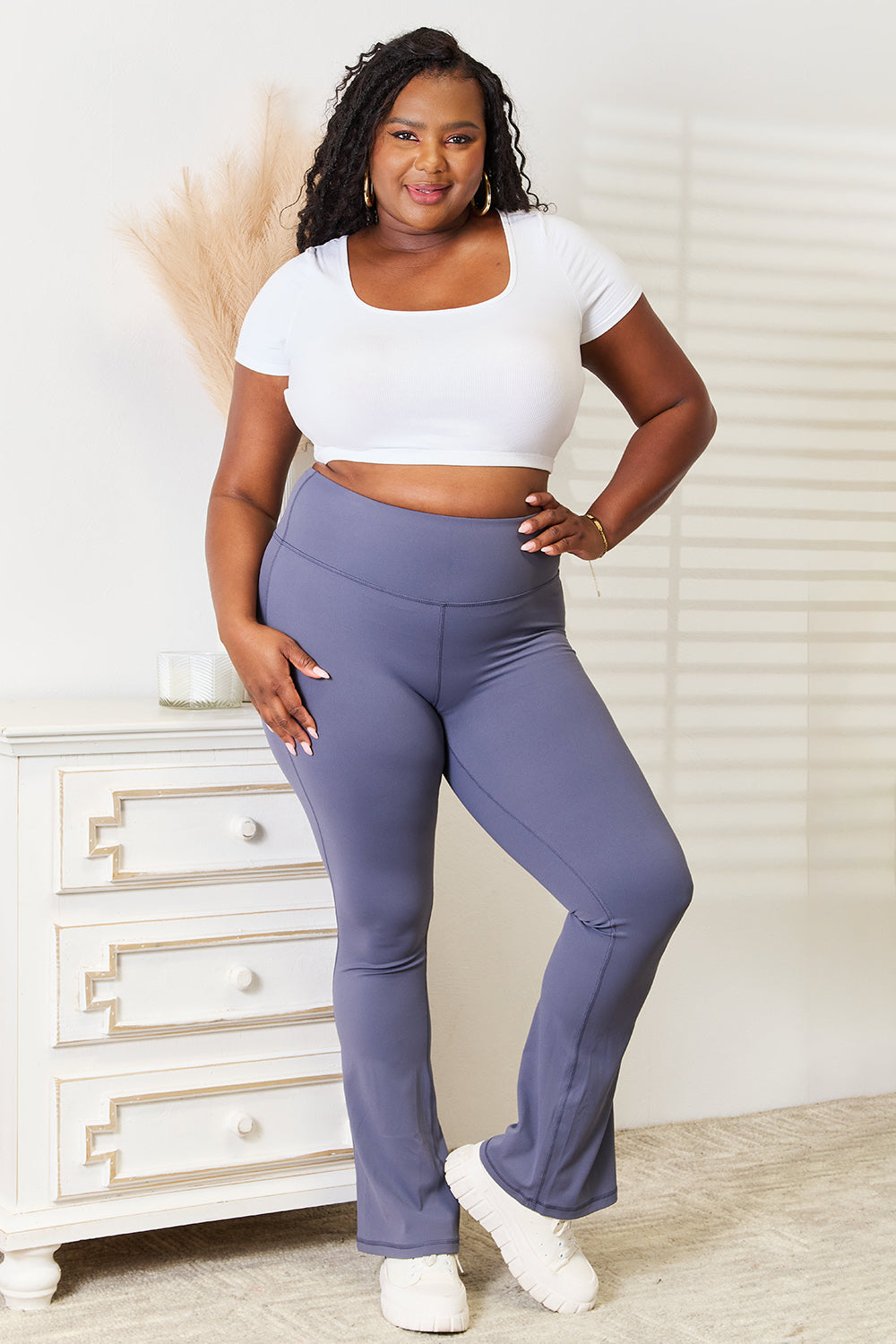 A person wearing a white crop top and the Millennia Basic Bae Wide Waistband Bootcut Sports Pants in high-waisted blue style stands indoors near a white dresser with gold accents.