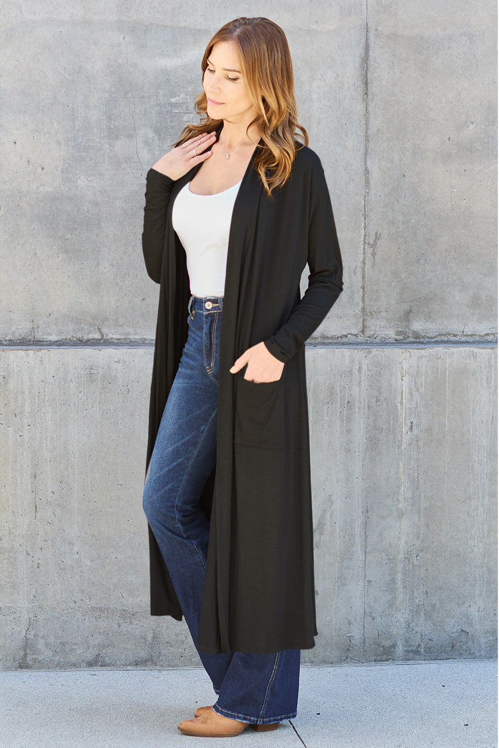 A woman stands against a concrete wall, wearing the Basic Bae Full Size Open Front Long Sleeve Cover Up made from rayon spandex, a white top, blue jeans in her perfect size, and brown shoes with her hands in her pockets. Machine wash cold for best results.