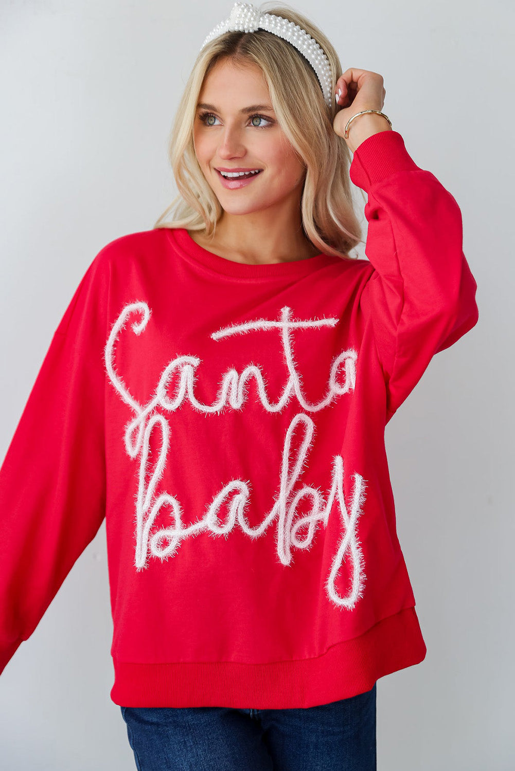 A person with long blonde hair is seen wearing the Fiery Red Christmas Santa Baby Tinsel Graphic Sweatshirt and blue jeans, facing away from the camera against a plain background.
