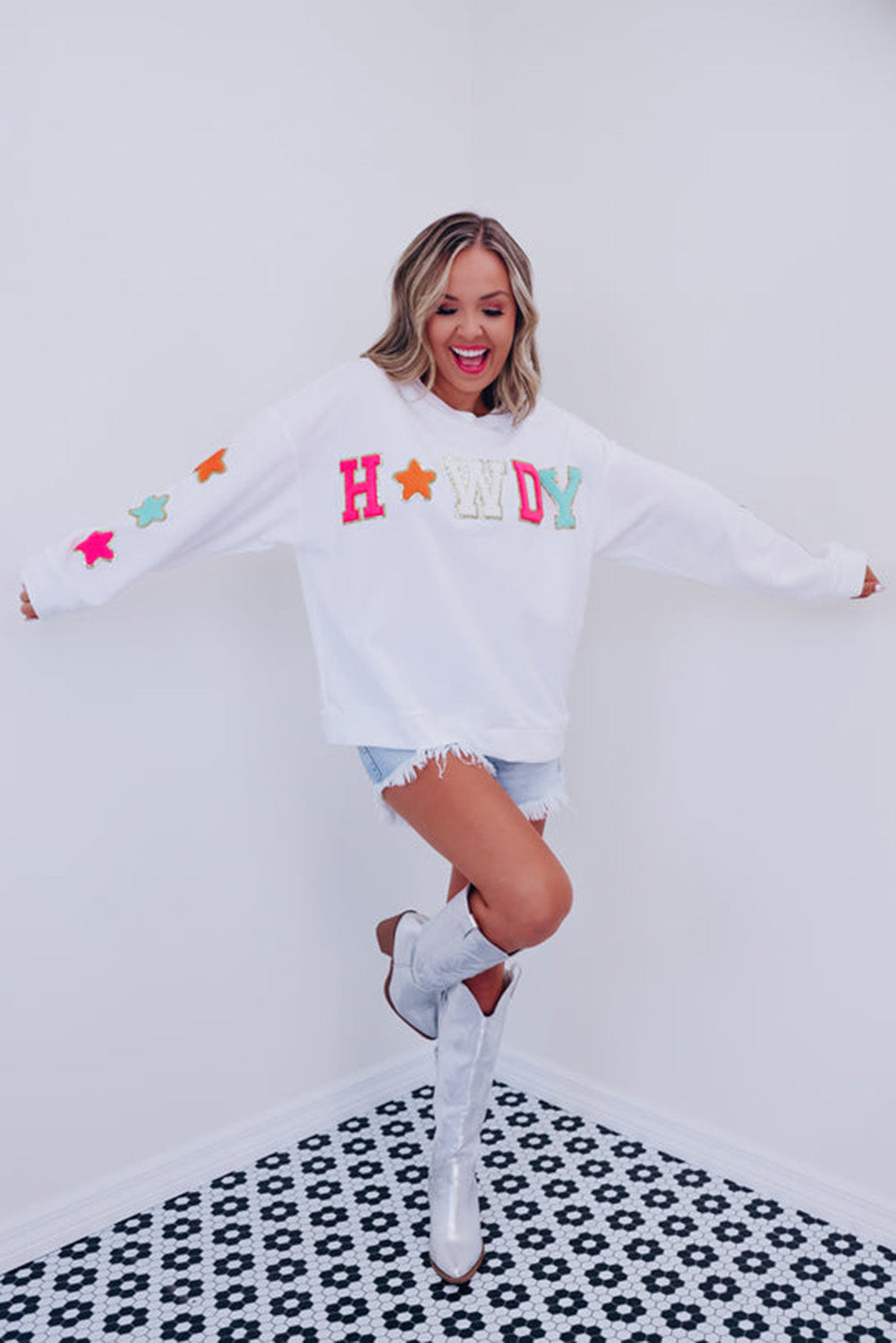 A person poses in the White Howdy Glitter Chenille Patch Graphic Casual Sweatshirt, adorned with colorful star patterns and the letters "WD." She wears light denim shorts and has shoulder-length blonde hair.