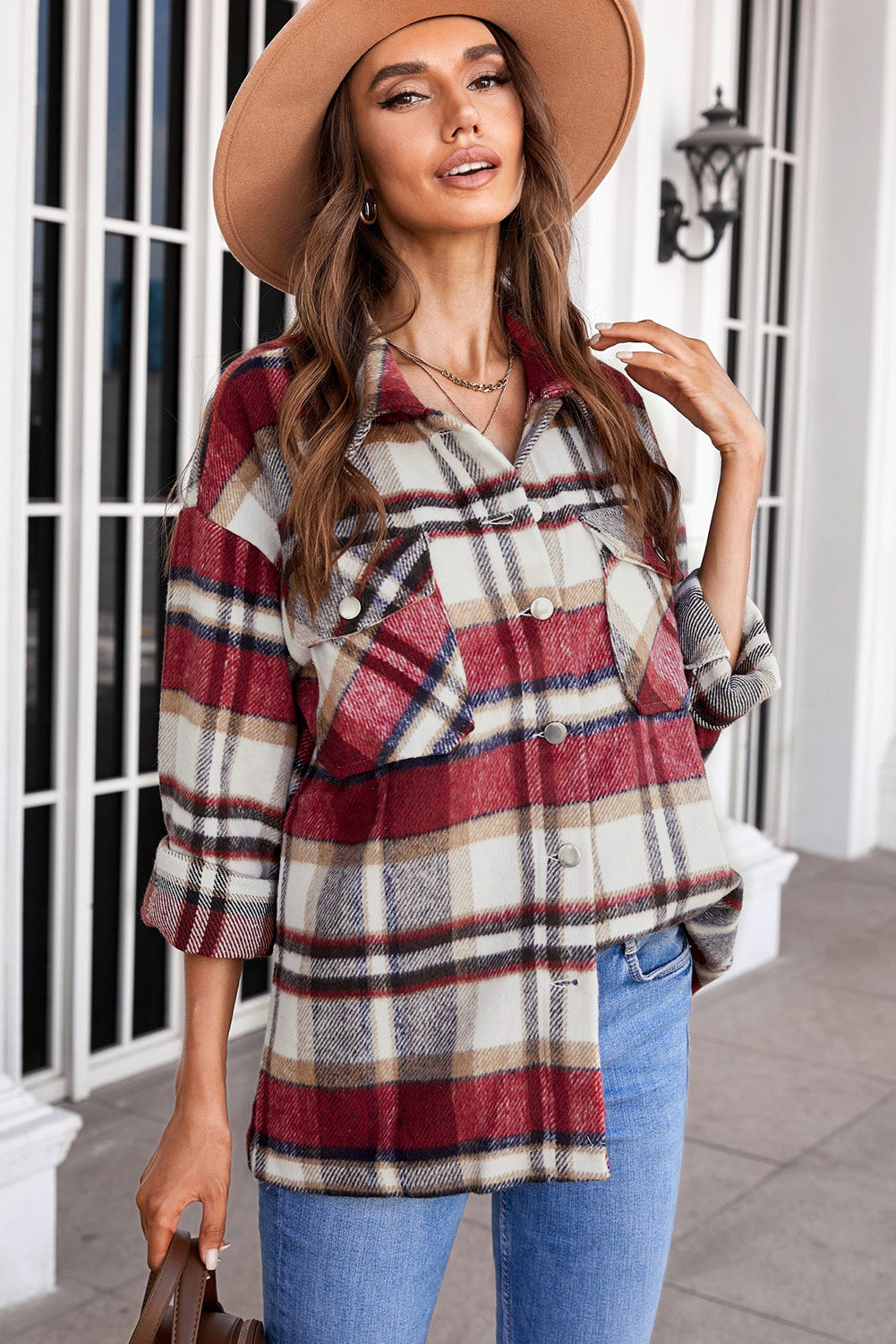 A person wearing a Brown Plaid Button Up Long Sleeve Flannel Shacket layered over a white top with ripped jeans and holding a brown handbag stands against a textured gray wall.