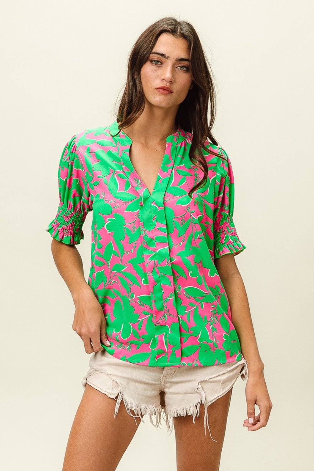 A woman wearing the BiBi Smocked Cuff Floral Contrast Top, featuring a chic design with bright pink and green floral patterns and ruffled sleeves, poses with one hand near her chin.