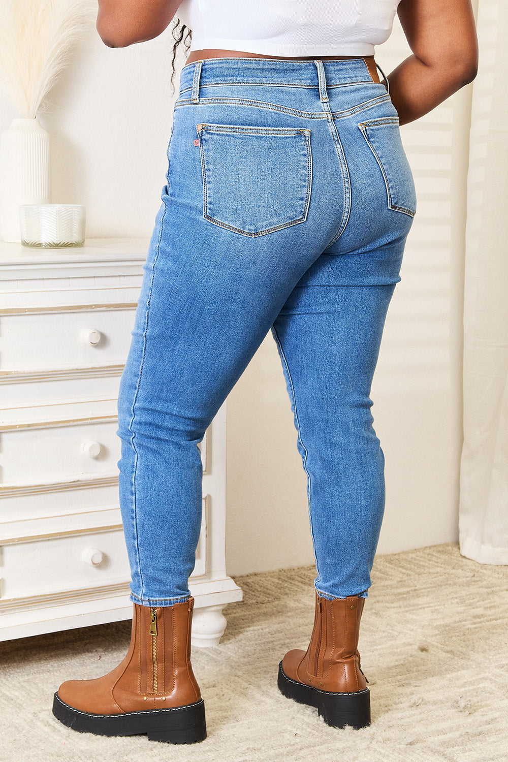 A person wearing Judy Blue Full Size High Waist Skinny Jeans in medium ash wash with a flattering fit, a white top, and brown boots stands near a white dresser, casually slipping one hand into their pocket.