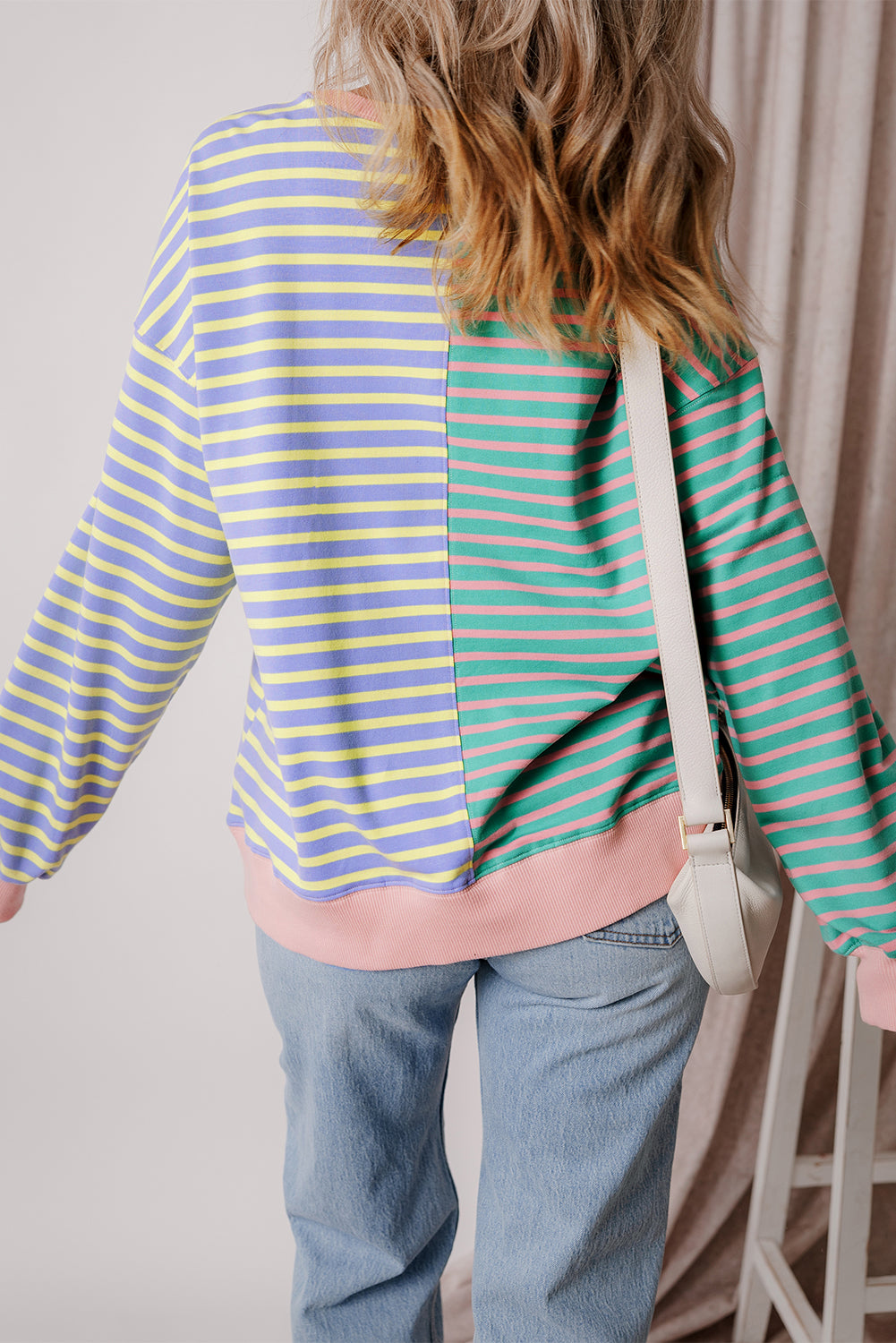A person with wavy hair is seen from behind wearing a Casual Stripe Colorblock Drop Shoulder Oversize Sweatshirt in pastel colors and blue jeans. They are carrying a white shoulder bag, showcasing a trendy color block design.