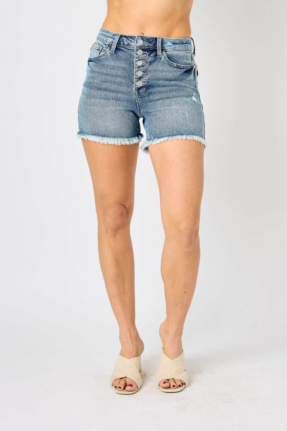 Wearing the Judy Blue Full Size Button Fly Raw Hem Denim Shorts and a white t-shirt, a person stands smiling in sandals against a plain background.