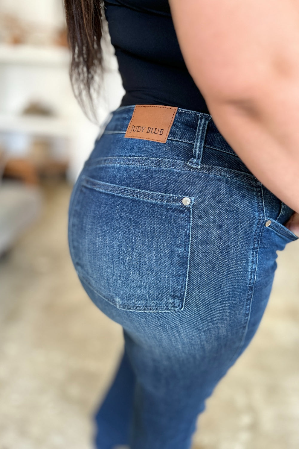 A person wearing a black top and Judy Blue Full Size Tummy Control Straight Jeans stands indoors with one hand on their hip. Their shoes are black with white soles. A plant and shelves are in the background.