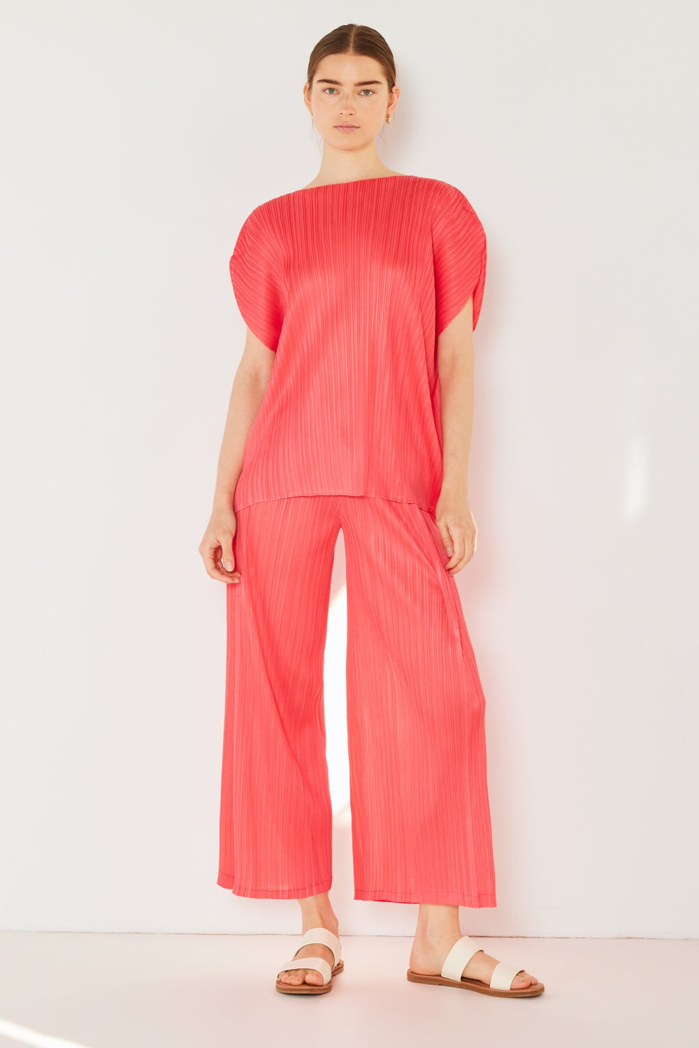 A person wearing the Marina West Swim Pleated Wide-Leg Pants with side pleat detail in red, standing against a white background with hands in pockets and wearing white sandals.