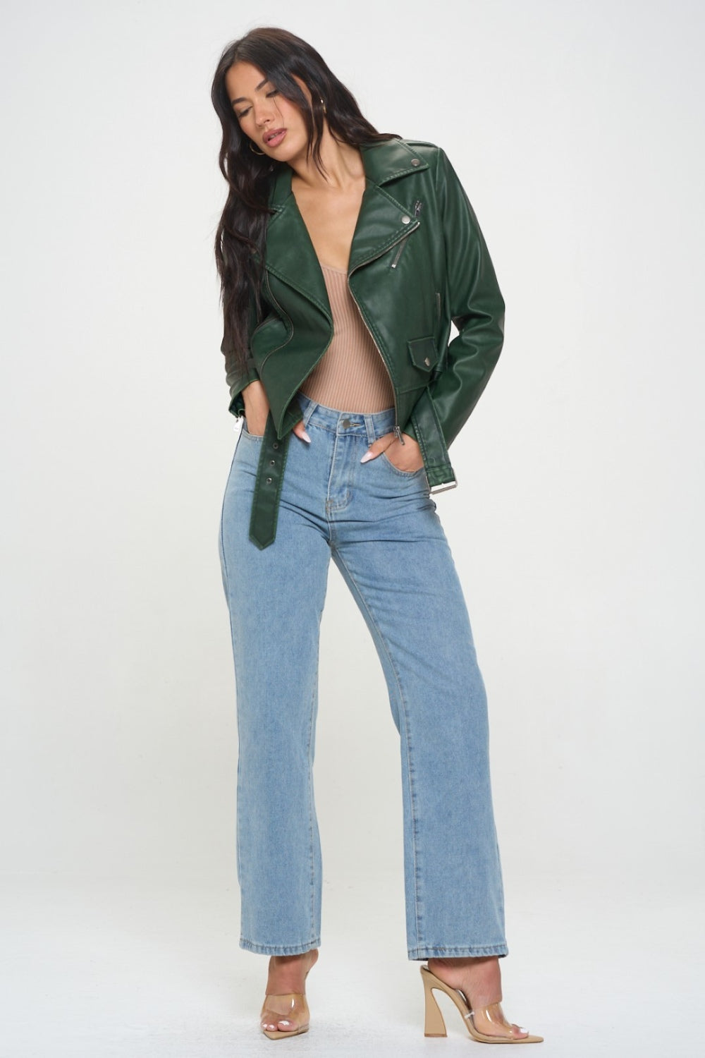 A person with long dark hair wearing a green Coalition LA Zip Up Biker Jacket with Belt and blue jeans stands against a plain background.