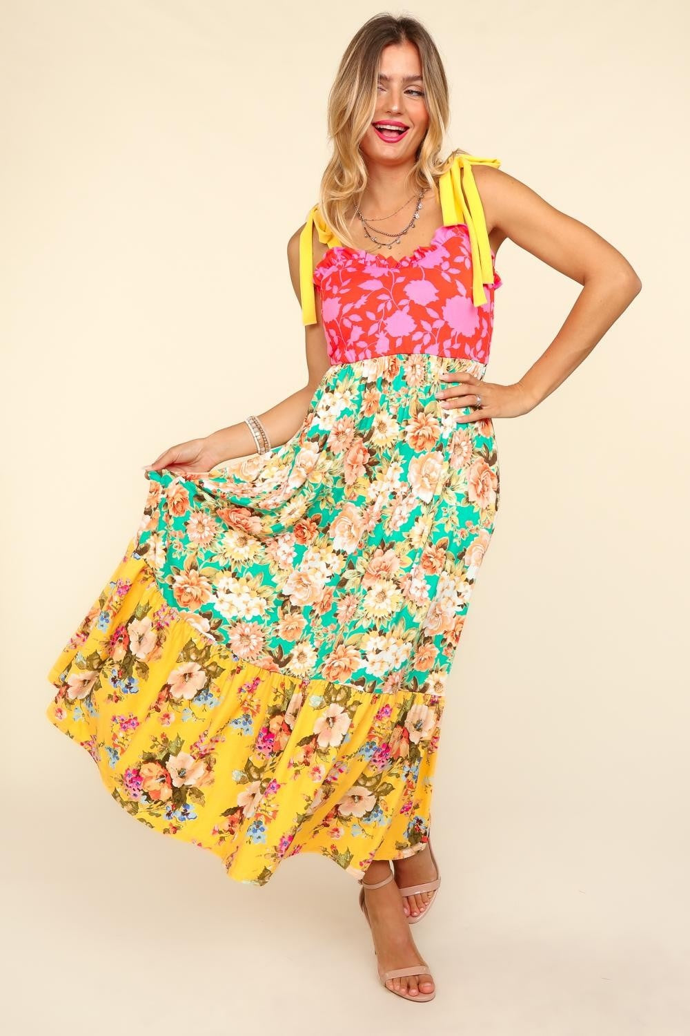 A person wearing the Haptics Floral Color Block Maxi Dress with Pockets, featuring a vibrant floral print and a color block maxi skirt in pink and yellow hues, stands smiling against a beige background.