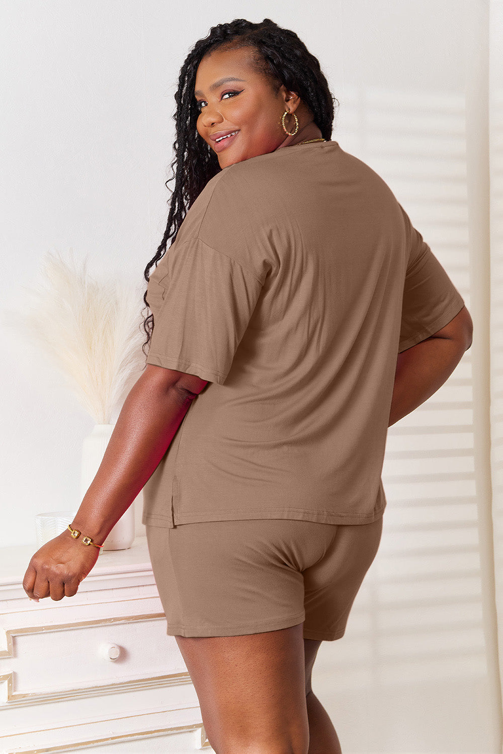 A woman is standing in a brightly lit room, wearing the Basic Bae Full Size Soft Rayon Half Sleeve Top and Shorts Set in a relaxed, stretchy turquoise fabric. She has one hand placed in her pocket and is smiling while looking slightly to the side.