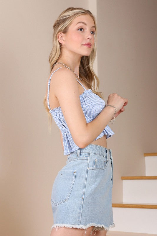 A woman wearing a Blue gingham tank top crafted from polyester spandex, paired with denim shorts, stands indoors and gazes to the side.