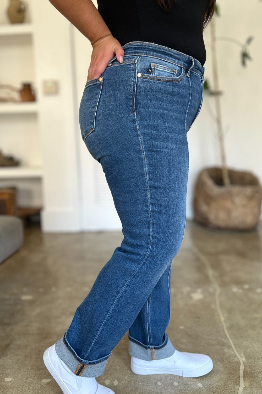 Two women stand side by side indoors, both wearing black tank tops, Judy Blue Full Size High Waist Front Seam Detail Straight Jeans, and white shoes. They are smiling and posing for the camera. A plant and shelves with decorations are in the background.