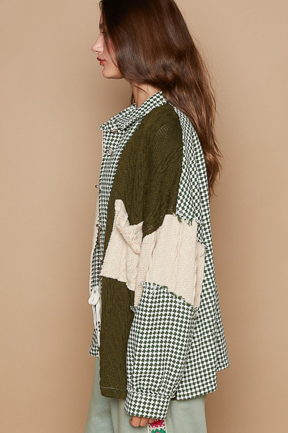 A person with curly hair is sitting on a wooden bench, wearing the POL Houndstooth Contrast Cable Knit Pocket Shacket over a light-colored top. Lush greenery is visible in the background.