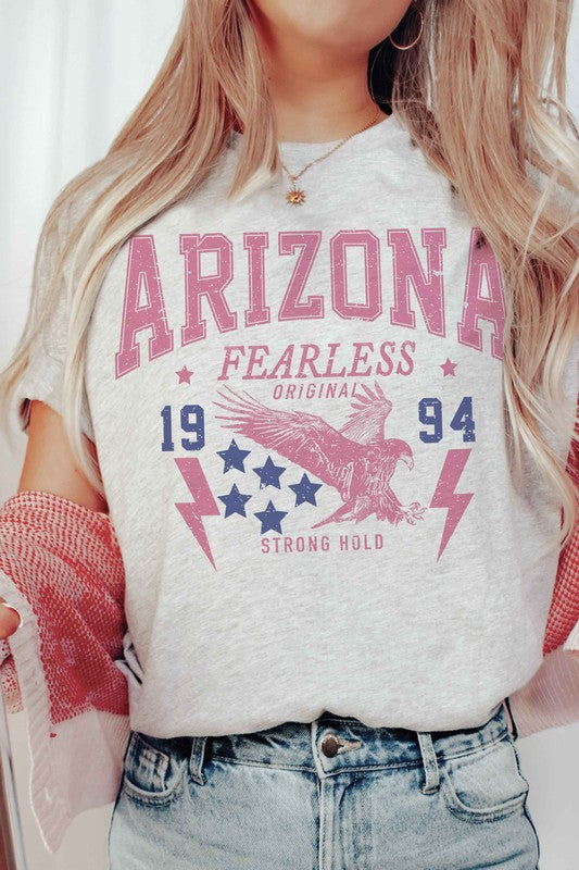 A woman with long light hair is wearing jeans and a 100% cotton ARIZONA EAGLE 1994 Graphic Tee, which features the text "ARIZONA FEARLESS ORIGINAL 1994 STRONG HOLD" along with an eagle, stars, and lightning bolts.