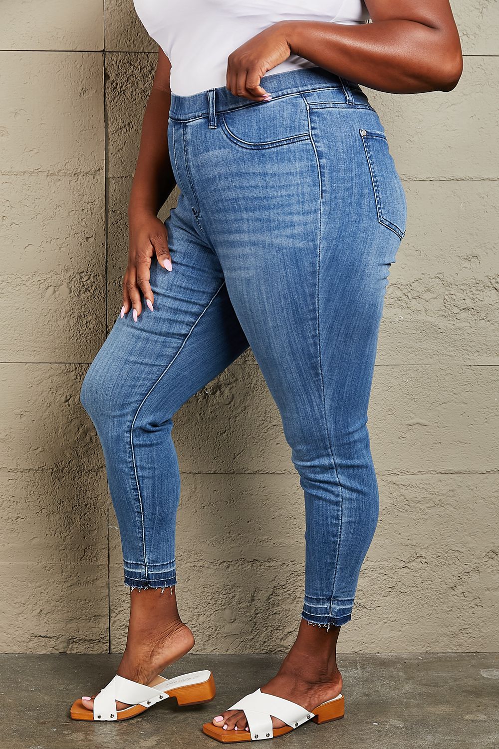 Person wearing Judy Blue Janavie Full Size High Waisted Pull On Skinny Jeans with release hem detailing and white sandals stands against a textured wall.