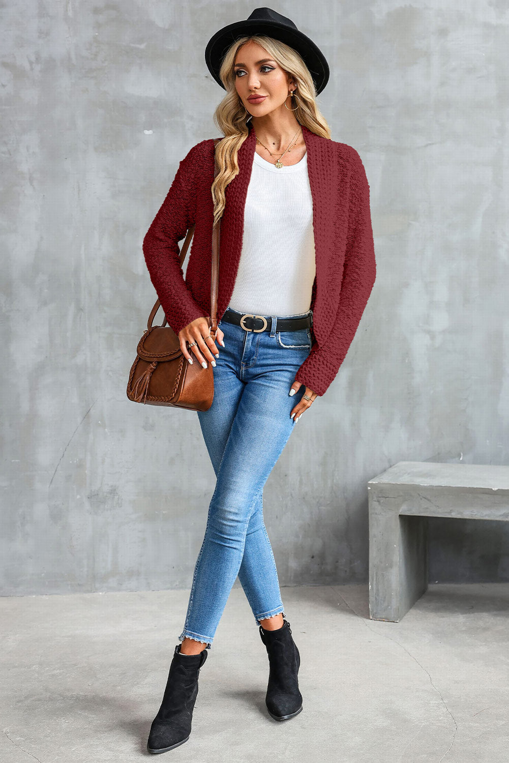 A woman in an Apricot Popcorn Knit Open Front Cardigan and jeans stands against a brick wall, looking to the side with a slight smile.