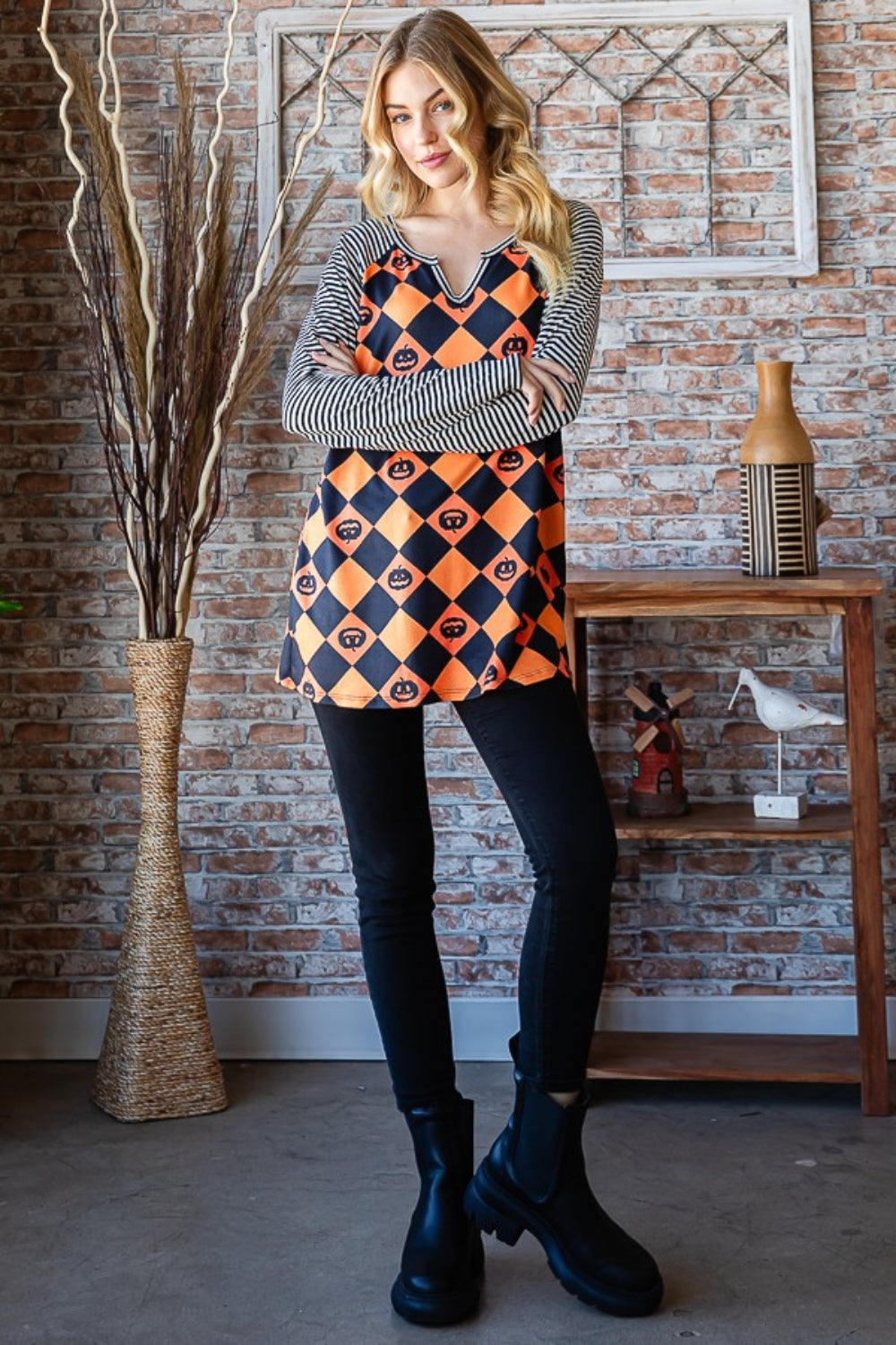 A person wearing the Heimish Full Size Checkered Contrast Striped Long Sleeve T-Shirt, which boasts a casual style with a black and orange checkered pattern adorned with pumpkin designs and contrasting black and white striped sleeves, stands before a brick wall enhanced by dried branches in the backdrop.