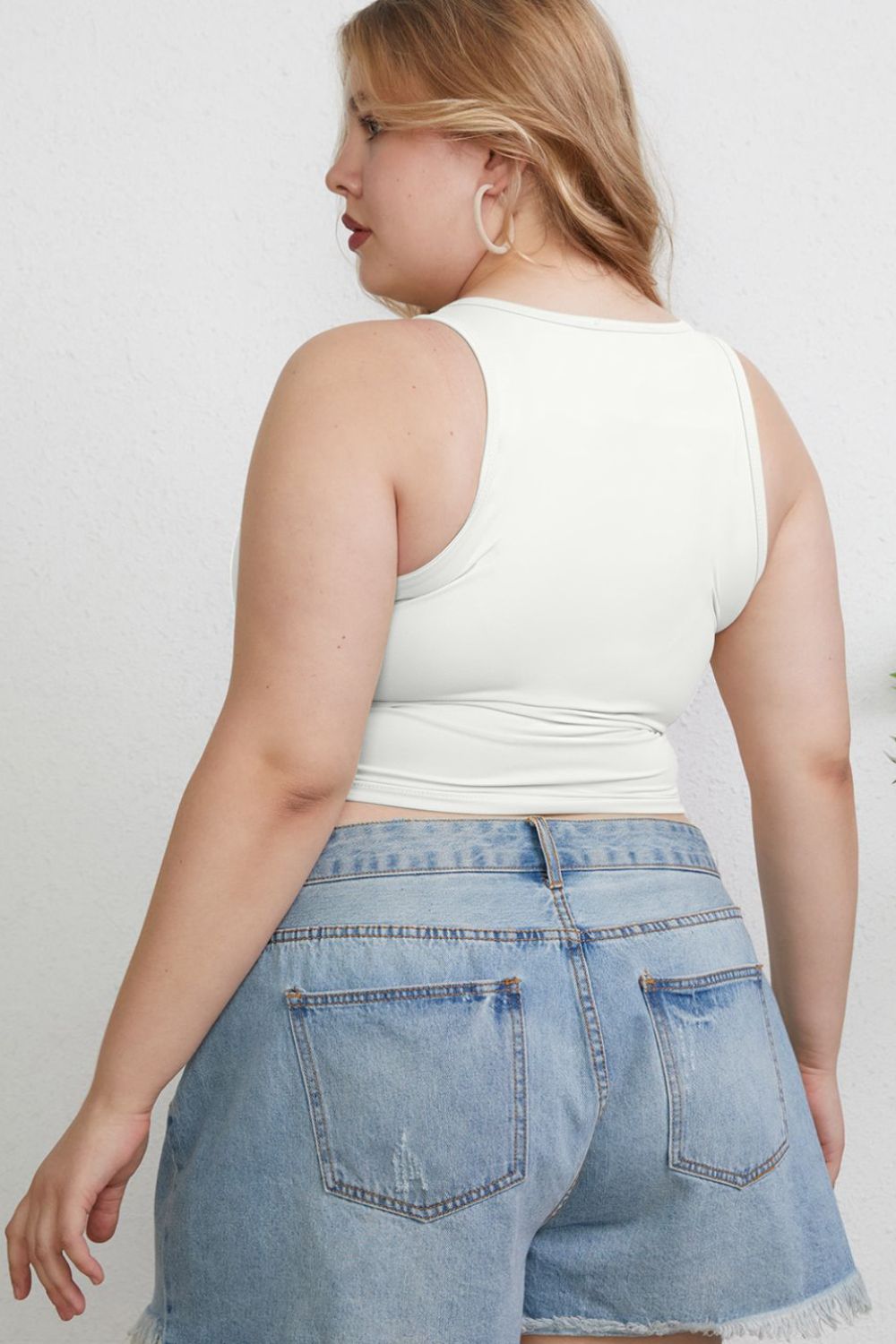 A woman with light hair wears the Basic Bae Full Size Round Neck Slim Tank in black and blue jeans, standing against a light-colored curtain.