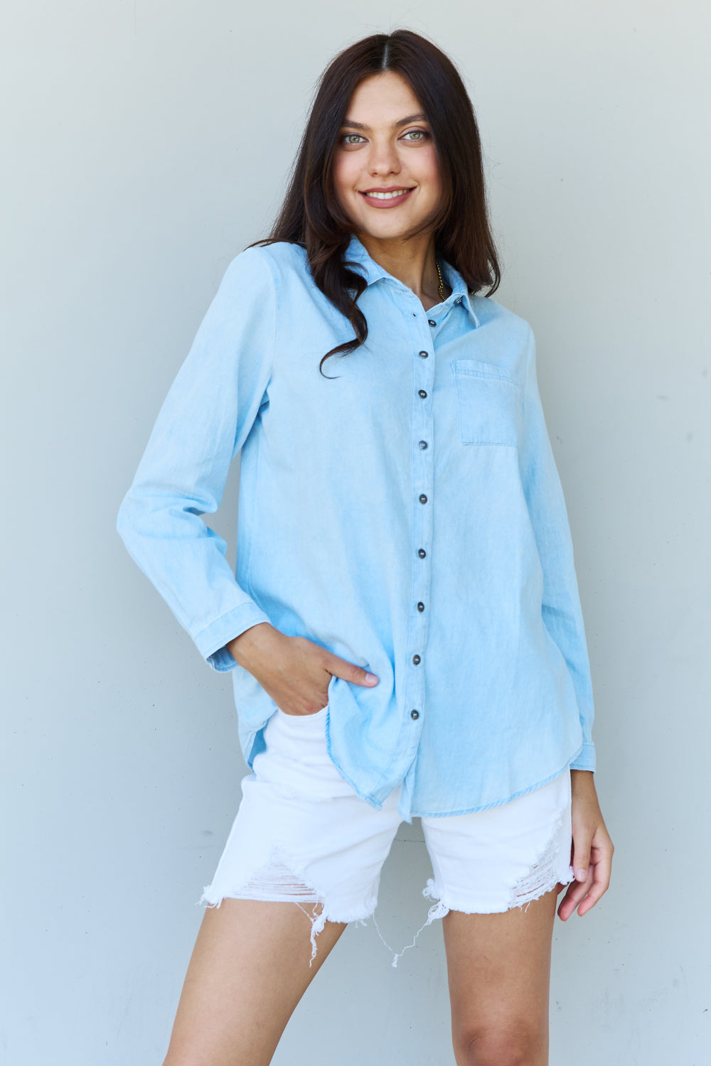 A woman wearing the Doublju Blue Jean Baby Denim Button Down Shirt Top in Light Blue, paired with white ripped shorts, strikes a pose with one hand in her pocket against a plain background, highlighting the classic button-up silhouette.