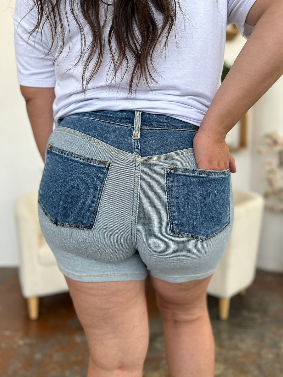 A person models indoors beside a beige chair and a plant, sporting a white t-shirt with the Judy Blue Full Size Color Block Denim Shorts for an effortlessly bold, eye-catching look.