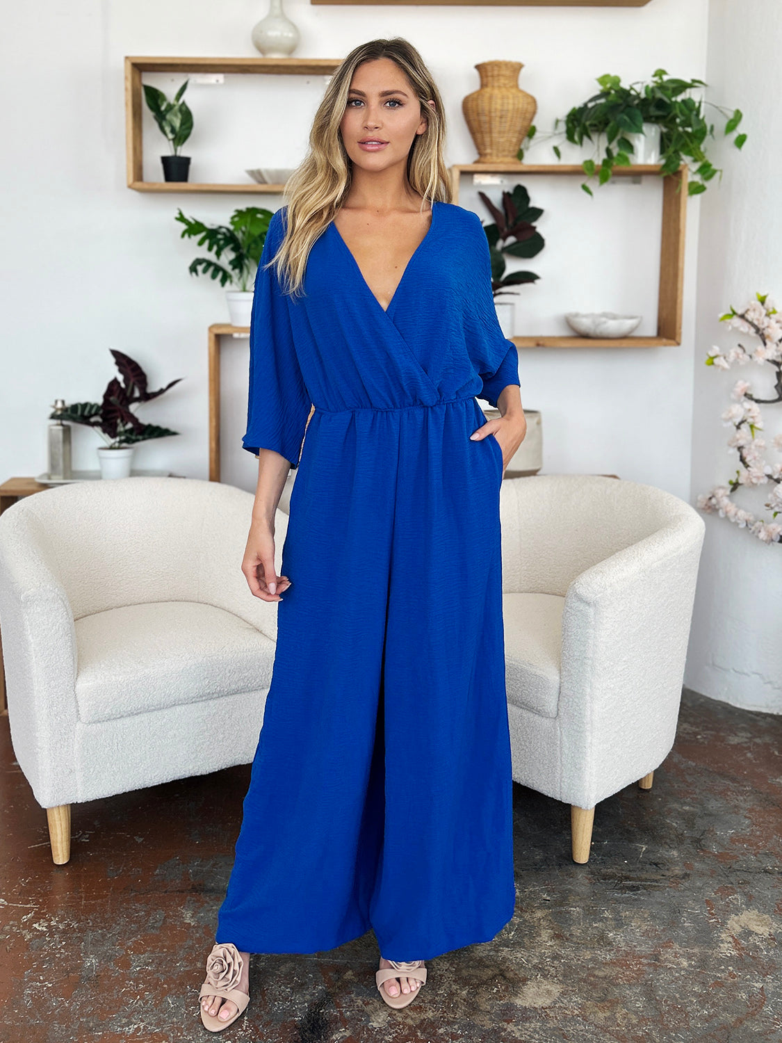 Person wearing the Double Take Full Size Surplice Wide Leg Jumpsuit with Pockets in royal blue, featuring wide legs, a deep V-neckline, and ruched detailing, posing with hands on hips against a plain light-colored background.