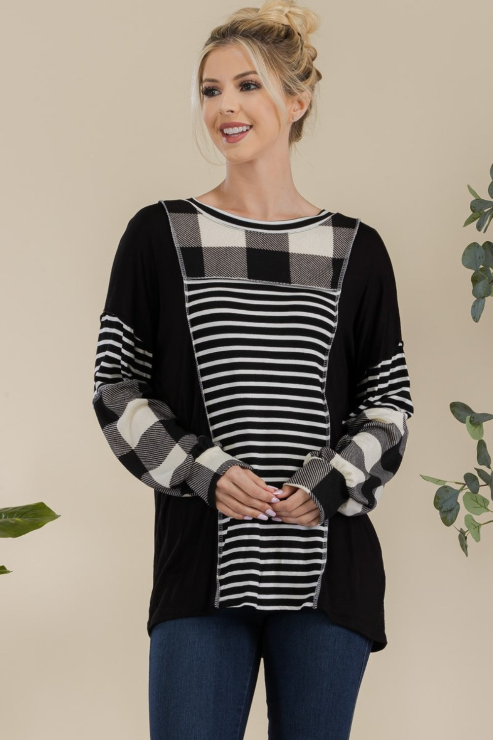 A person wearing a Celeste Full Size Color Block Exposed Seam T-Shirt, featuring black and white plaid with reverse stitching and long sleeves, poses while smiling in front of a neutral background with a plant.