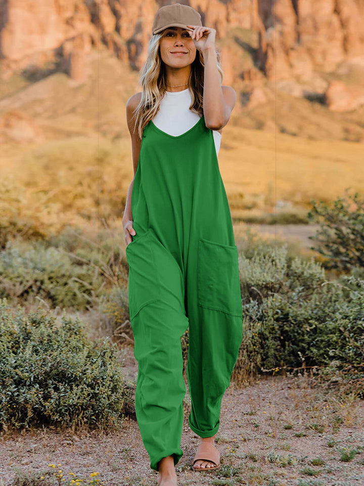 A woman wearing the Double Take Full Size Sleeveless V-Neck Pocketed Jumpsuit in green and a beige cap stands outdoors in a desert landscape with plants and rocky cliffs in the background.