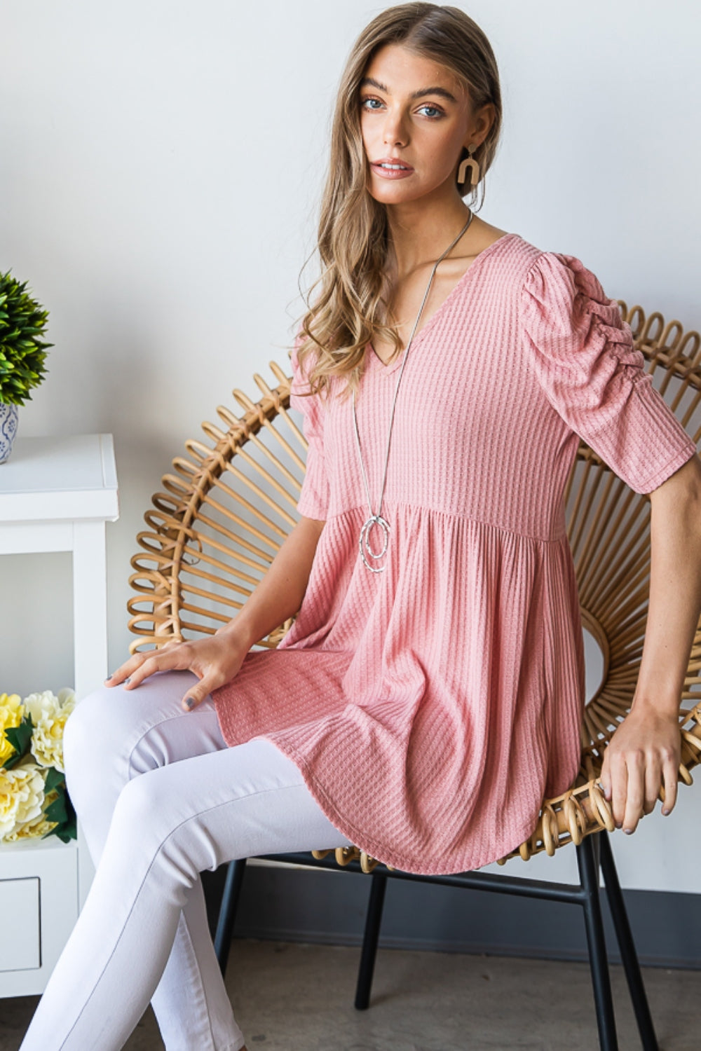 A woman in a Heimish Full Size Waffle Knit V-Neck Babydoll Top with short-sleeves and a long necklace, standing by a window with a neutral expression; the perfect cozy wardrobe addition.