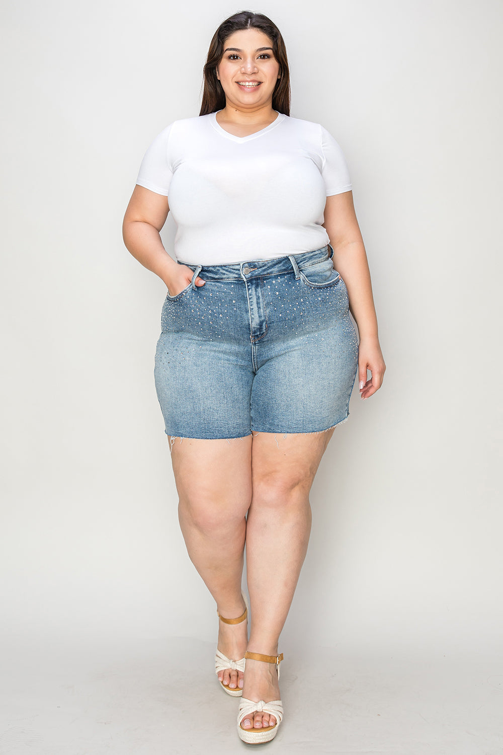 A woman wearing a white T-shirt, Judy Blue Full Size High Waist Rhinestone Decor Denim Shorts, and white sandals poses against a plain background.