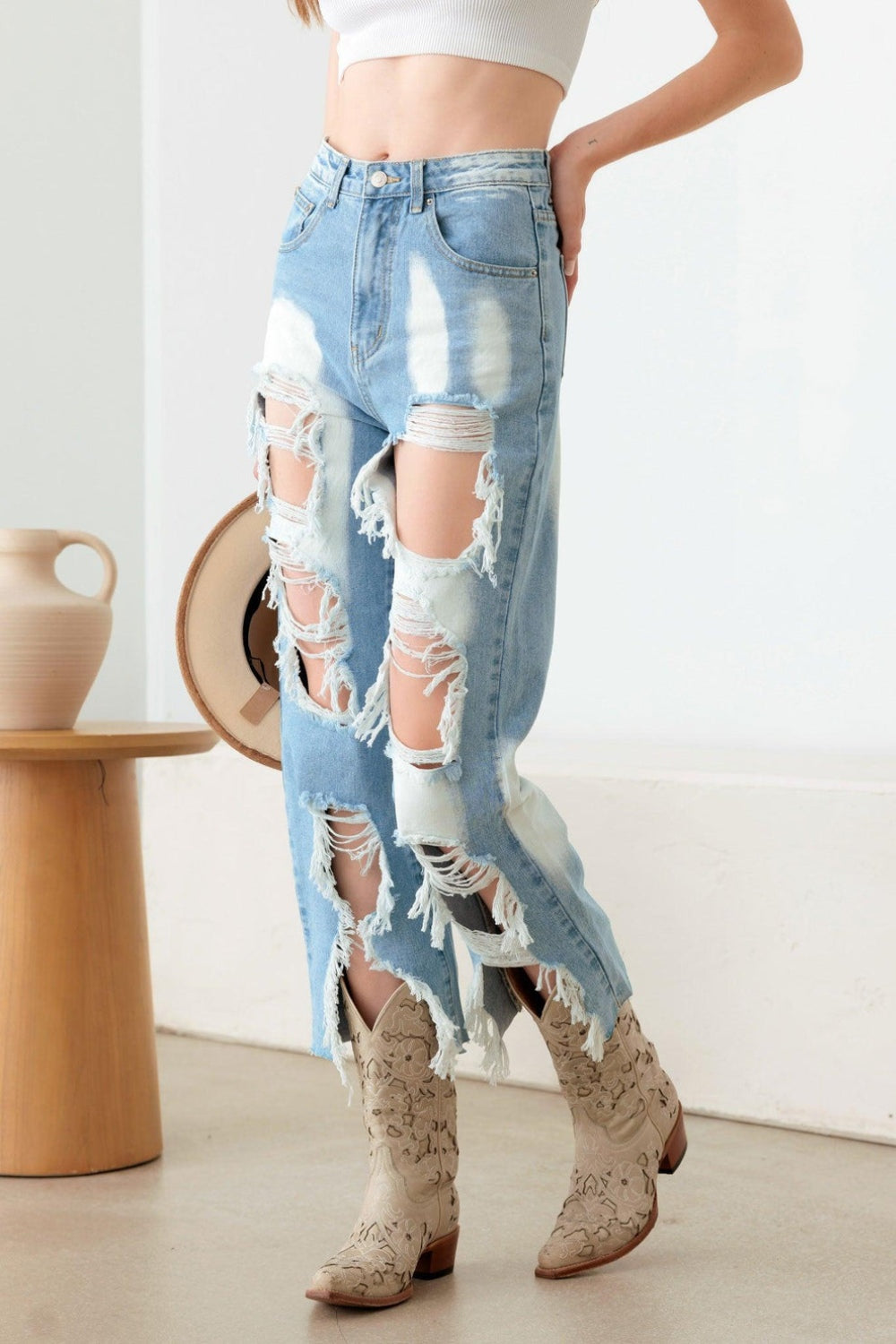 A woman stands indoors in a Western Boho style, showcasing a white crop top and plaid shirt paired with the Litz La Frayed Cut Distressed Jeans. She holds a hat in her hand and completes her outfit with patterned boots, while a vase is positioned on a wooden stool nearby.