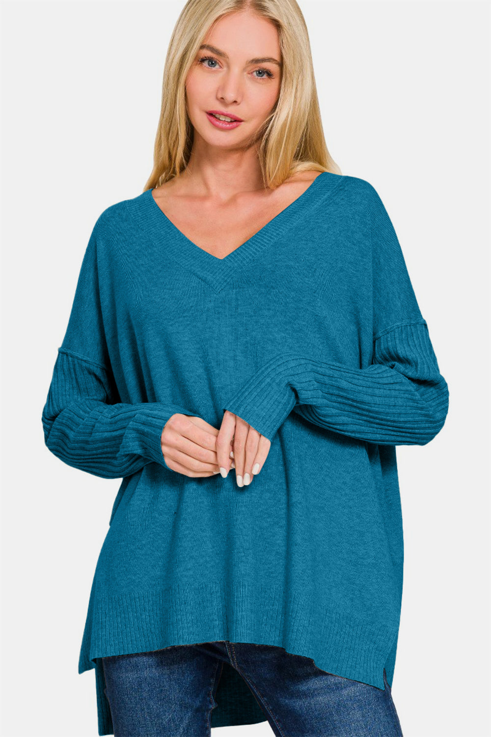 A woman with long blonde hair wears a Zenana V-Neck Side Slit High-Low Sweater in teal, featuring ribbed sleeves, alongside blue jeans, and stands against a plain white background.