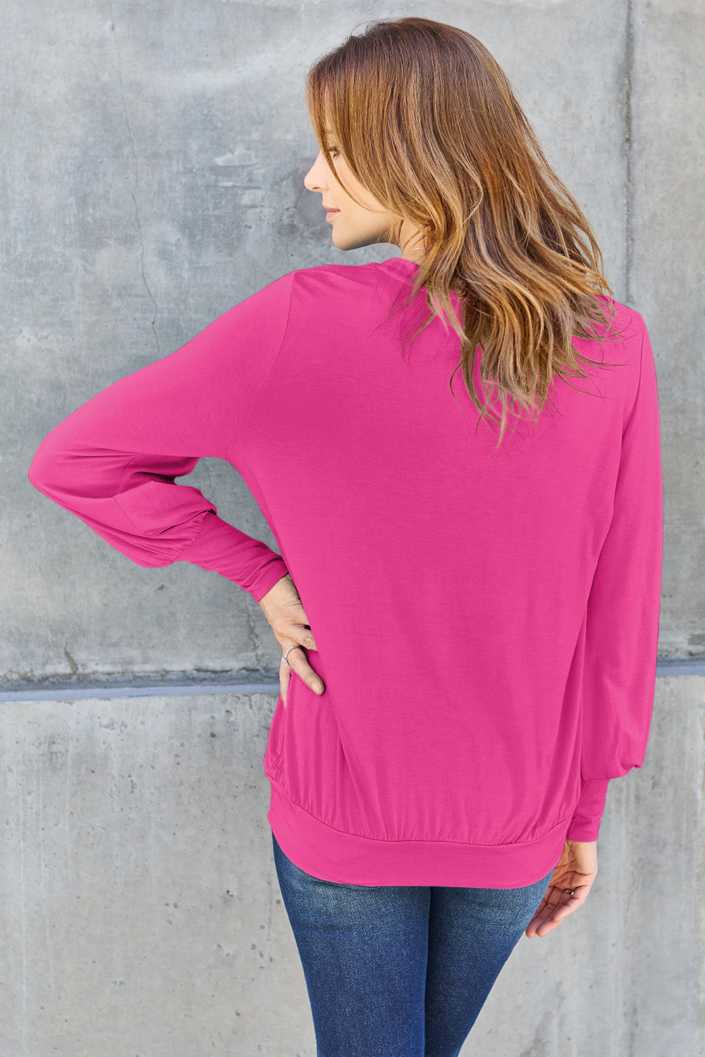 A woman with brown hair is standing against a concrete wall, wearing the Basic Bae Full Size V-Neck Lantern Sleeve Top in teal and blue basic-style jeans, smiling.