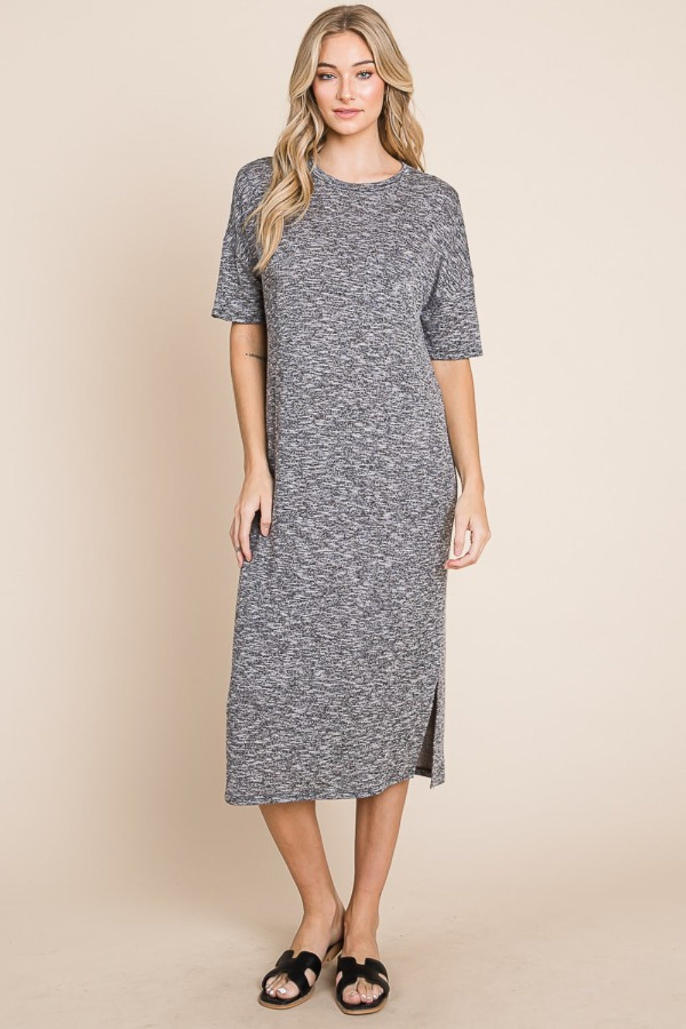 A woman wearing the BOMBOM Slit Round Neck Half Sleeve Midi Dress, a knee-length, grey midi dress featuring short sleeves and a slit detail at the hem, paired with black slip-on sandals, stands in front of a neutral background.