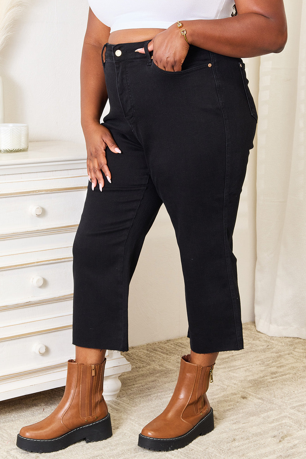 A person wearing Judy Blue Full Size High Waist Wide Raw Hem Cropped Jeans in black and a white lace top stands next to a white dresser adorned with a vase and candle.