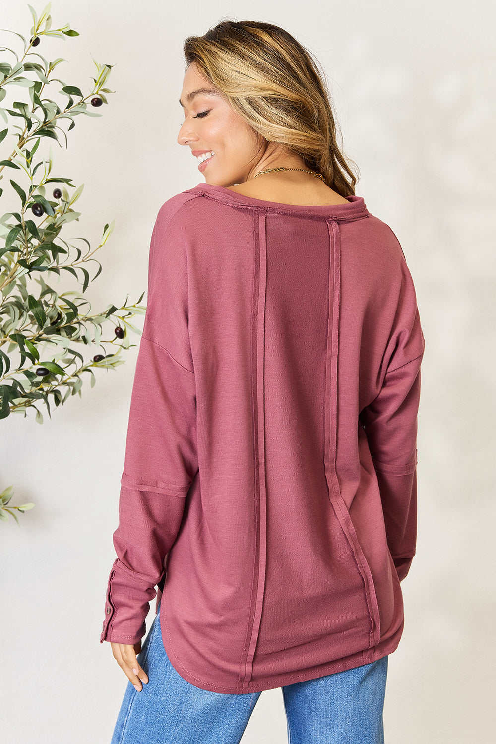 A person stands in front of a plain backdrop with a plant, wearing a maroon Culture Code Full Size V-Neck Exposed Seam Long Sleeve Blouse and blue jeans, smiling slightly with their hand on their hip. The relaxed fit of the polyester blend adds to the casual yet stylish look.