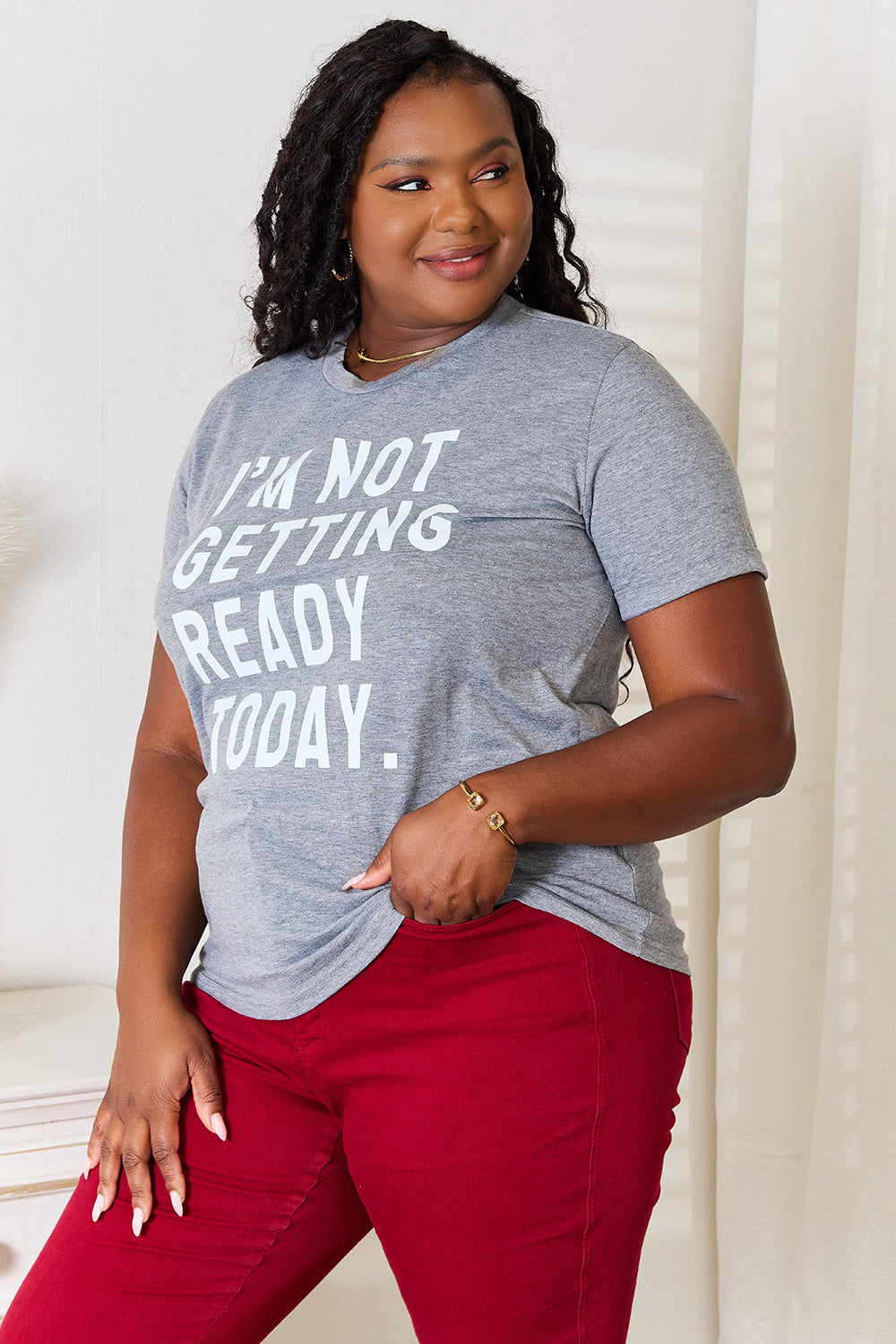 Dressed in a Simply Love I'M NOT GETTING READY TODAY Graphic T-Shirt and vibrant red pants, the individual exudes a carefree style while standing near a white surface adorned with decorative objects.