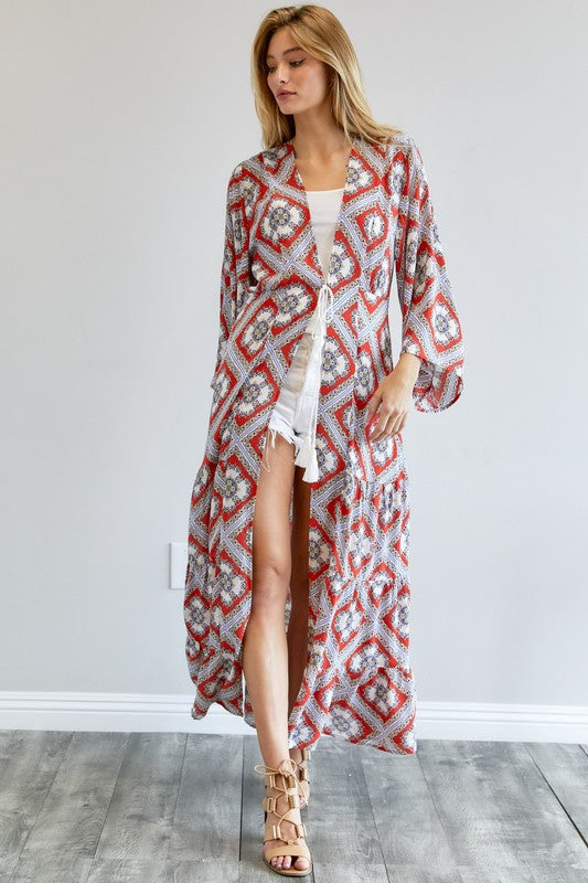 A person wearing a Printed Long Sleeve Loose Kimono with a bohemian blue and pink print over a white outfit stands in a room with a gray floor and white wall.