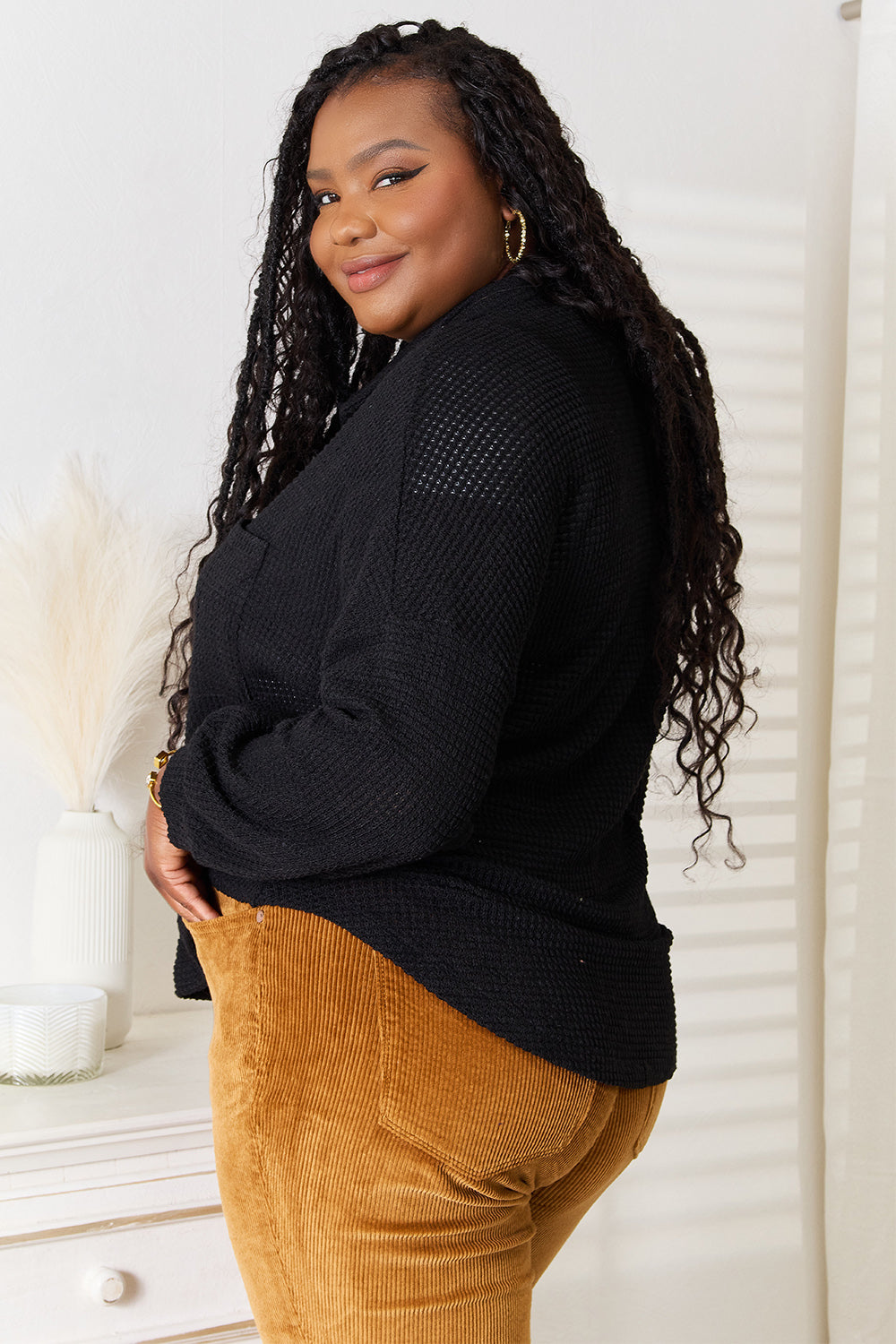 A woman with long hair is wearing a Double Take Waffle-Knit Collared Neck Dropped Shoulder Shirt in black and mustard-colored pants. She stands indoors, resting one hand in her pocket and the other touching her hair.