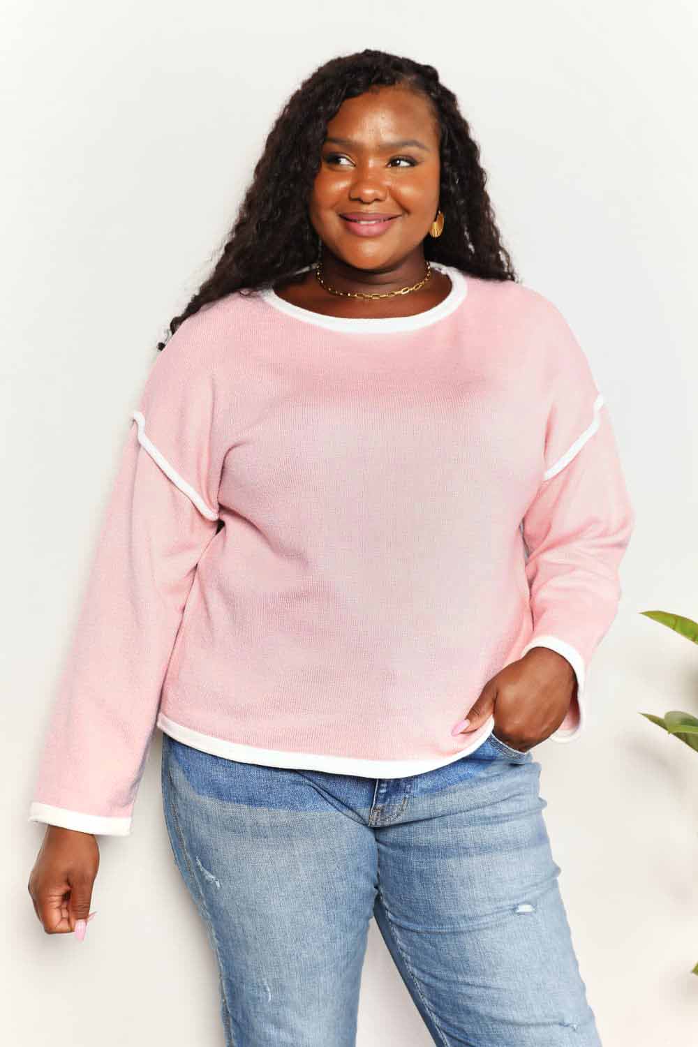 A woman with long curly hair is wearing the Angel Wings Contrast Detail Dropped Shoulder Knit Top in pink, paired with blue jeans, standing against a light background.