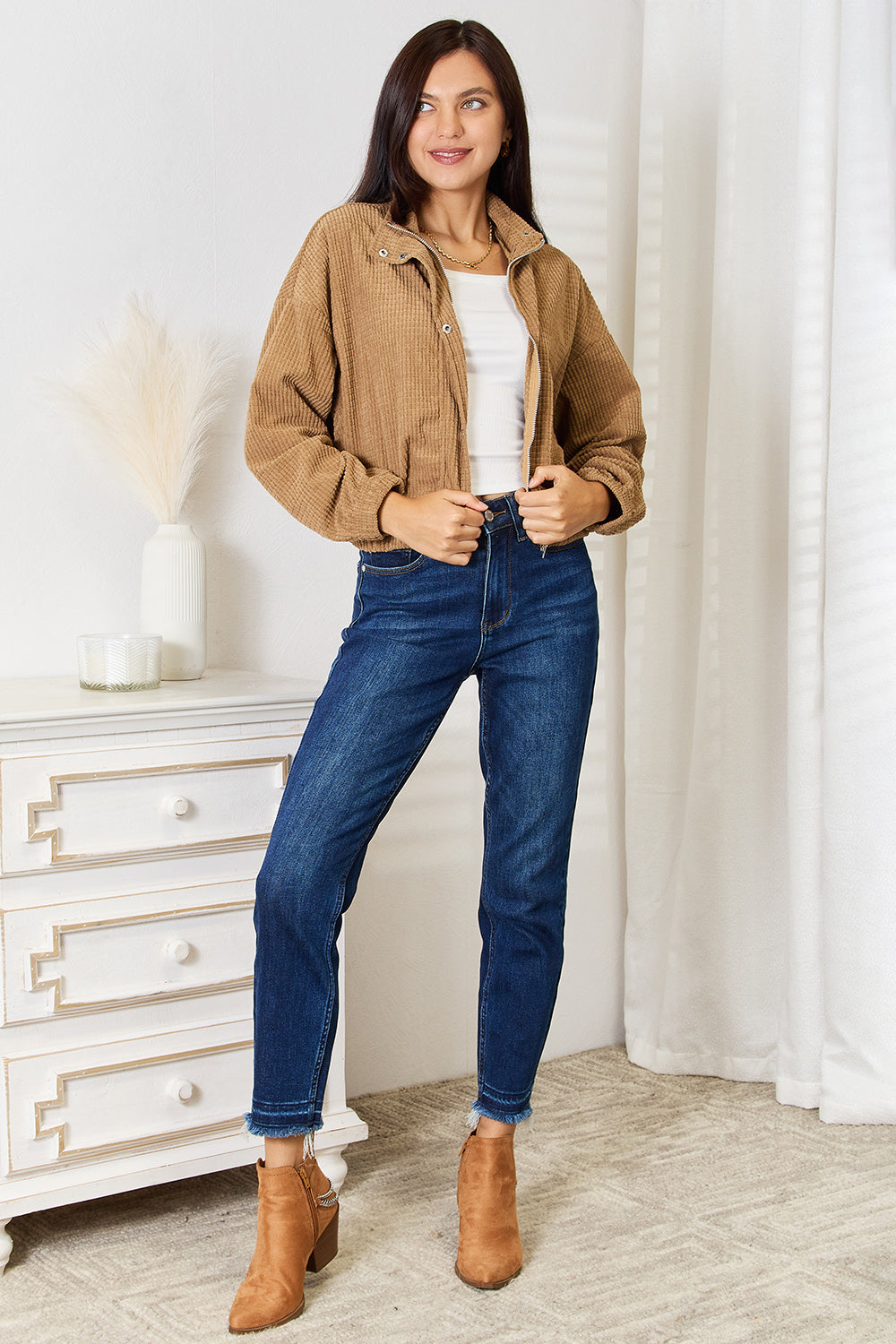 A person wearing the versatile Perfee Long Sleeve Dropped Shoulder Jacket in brown corduroy and blue jeans is standing indoors, smiling, with one hand resting on a white piece of furniture.